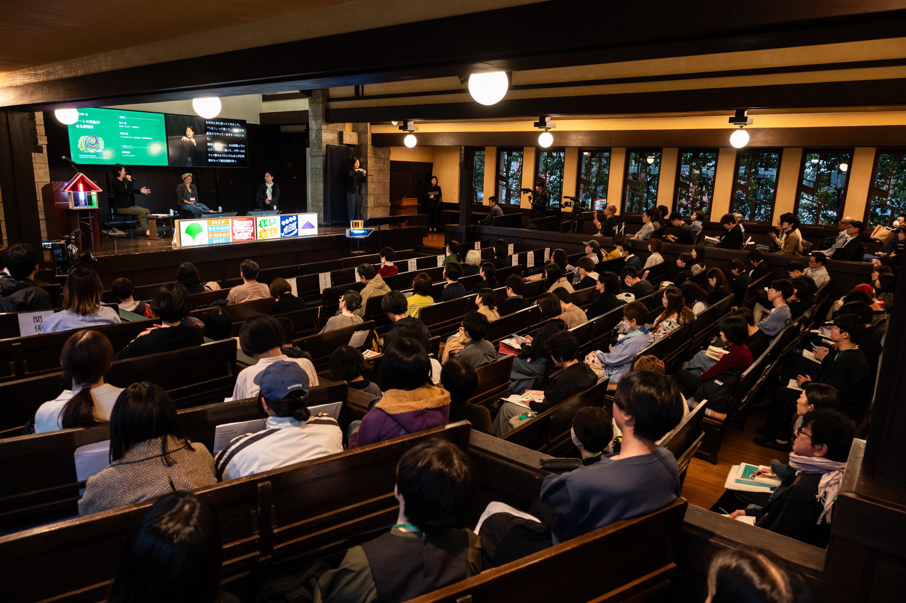 講堂内で、大勢の参加者が客席に座り、舞台上の3名の登壇者のセッションを聞いている様子。