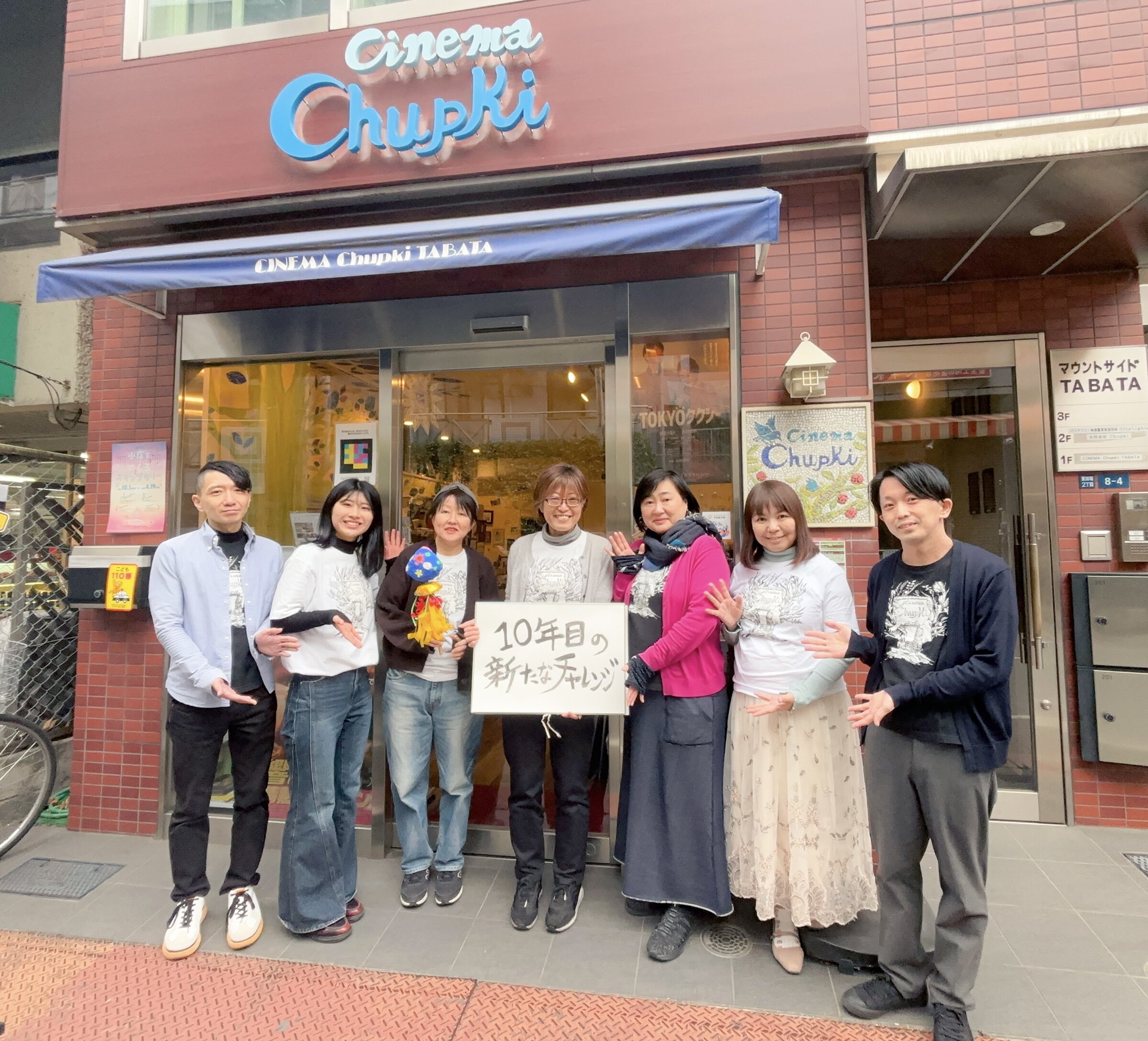 合同会社チュプキで働くスタッフ7人が自分たちの活動場所の建物の前で、「10年目の新たなチャレンジ」という看板をもって集合している写真