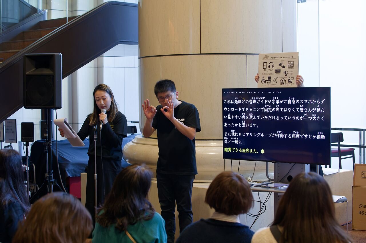 イベント中、参加者に向かって1人の女性がマイクを持って話をしていて、となりには手話をしている男性が立っている。