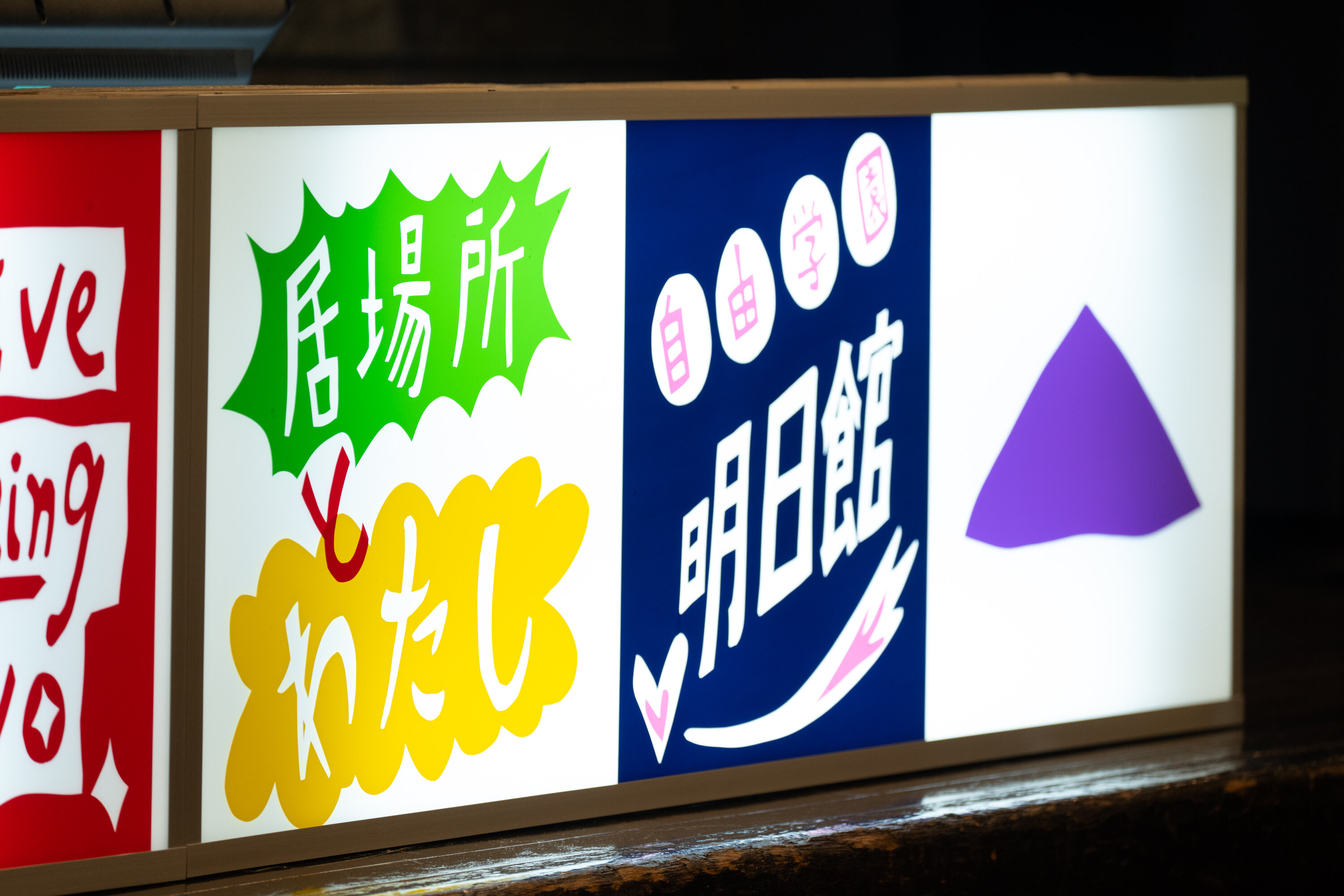 横長の看板作品の写真。看板はネオンの明かりのように発光している。写真左側に「居場所とわたし」、中央に「自由学園明日館」と手書きのような字でポップな感じで書かれている。右側には紫色の三角のマークが描かれている。