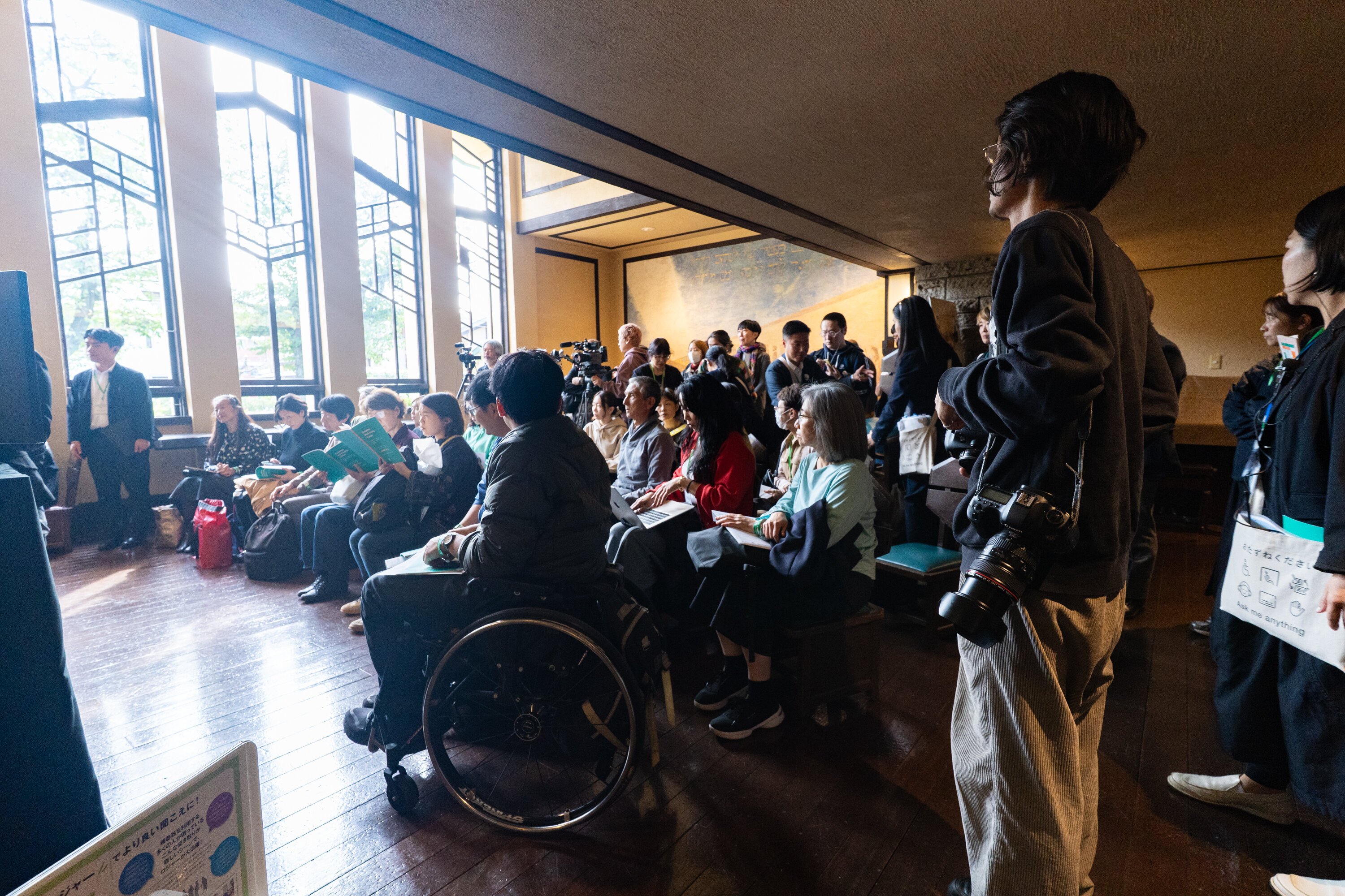自由学園ホールの観客の様子。正面奥にあるステンドグラスから光が差し込んでいる。２０名ほどの参加者が半円状の形で左に向かっている。手前には車いすに乗っている参加者もいる。