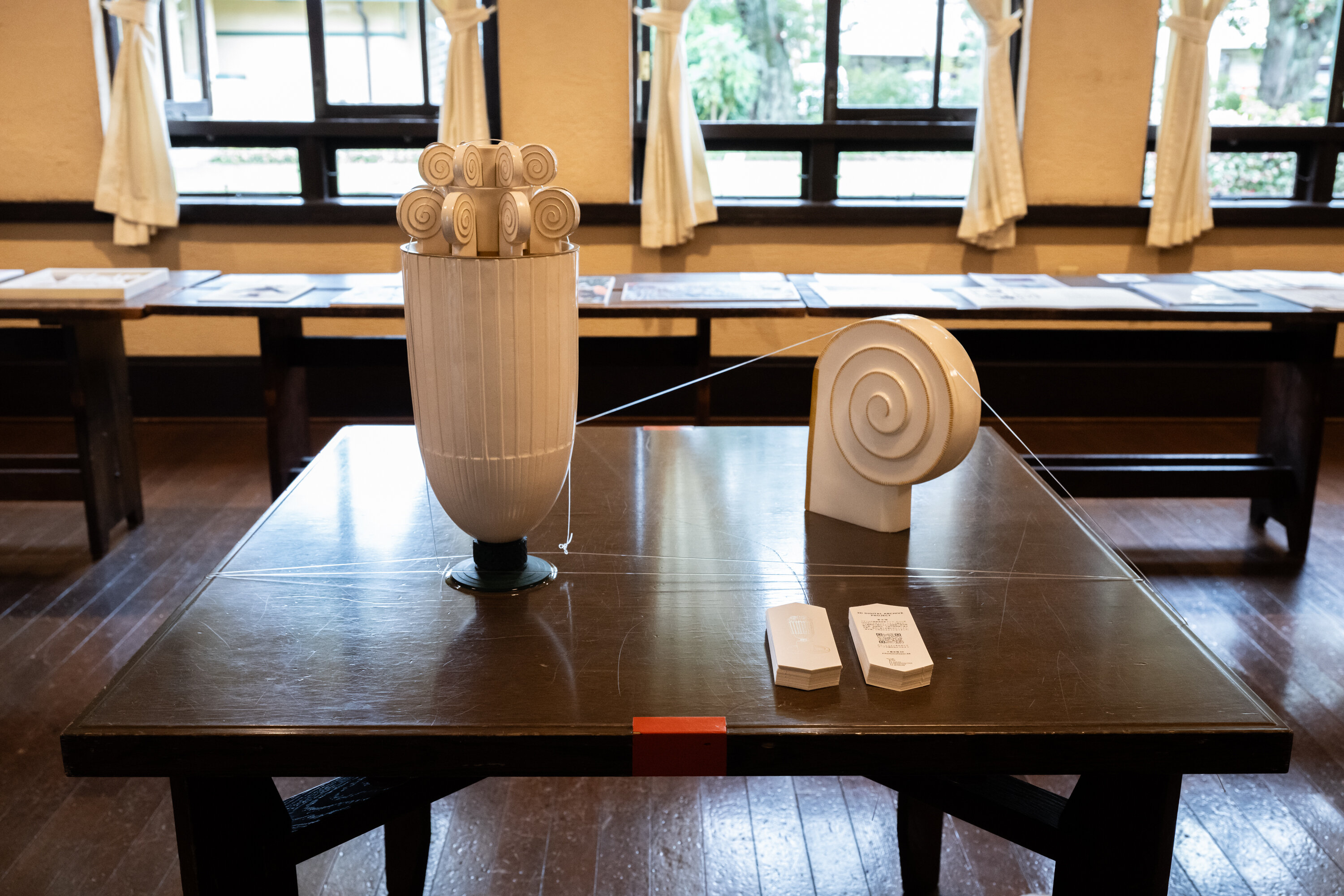 正方形の机の上に、白く大きなオブジェが2つ置かれている。左側のオブジェはパフェグラスのような形で、上部には渦巻き状の飾りが円状に並べられている。右側にはその渦巻き状の飾りを拡大したオブジェが置かれている。手前には説明カードが２つ、積まれて置かれている。