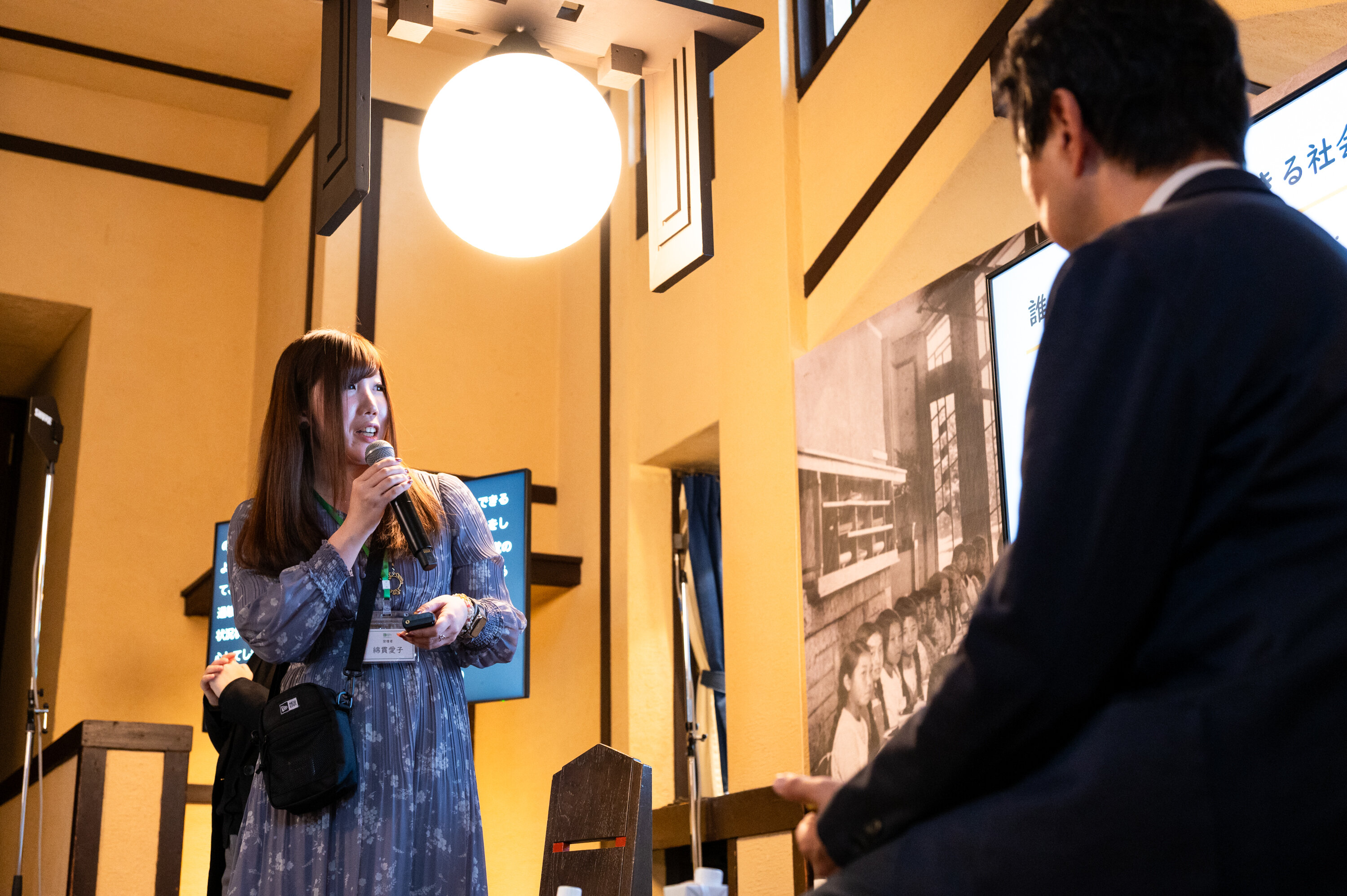 茶色の長髪でワンピースを着た登壇者がマイクを持ち、モニターのスライドを見ながら話している。手前にいるスーツ姿の登壇者がその説明を聞いている。