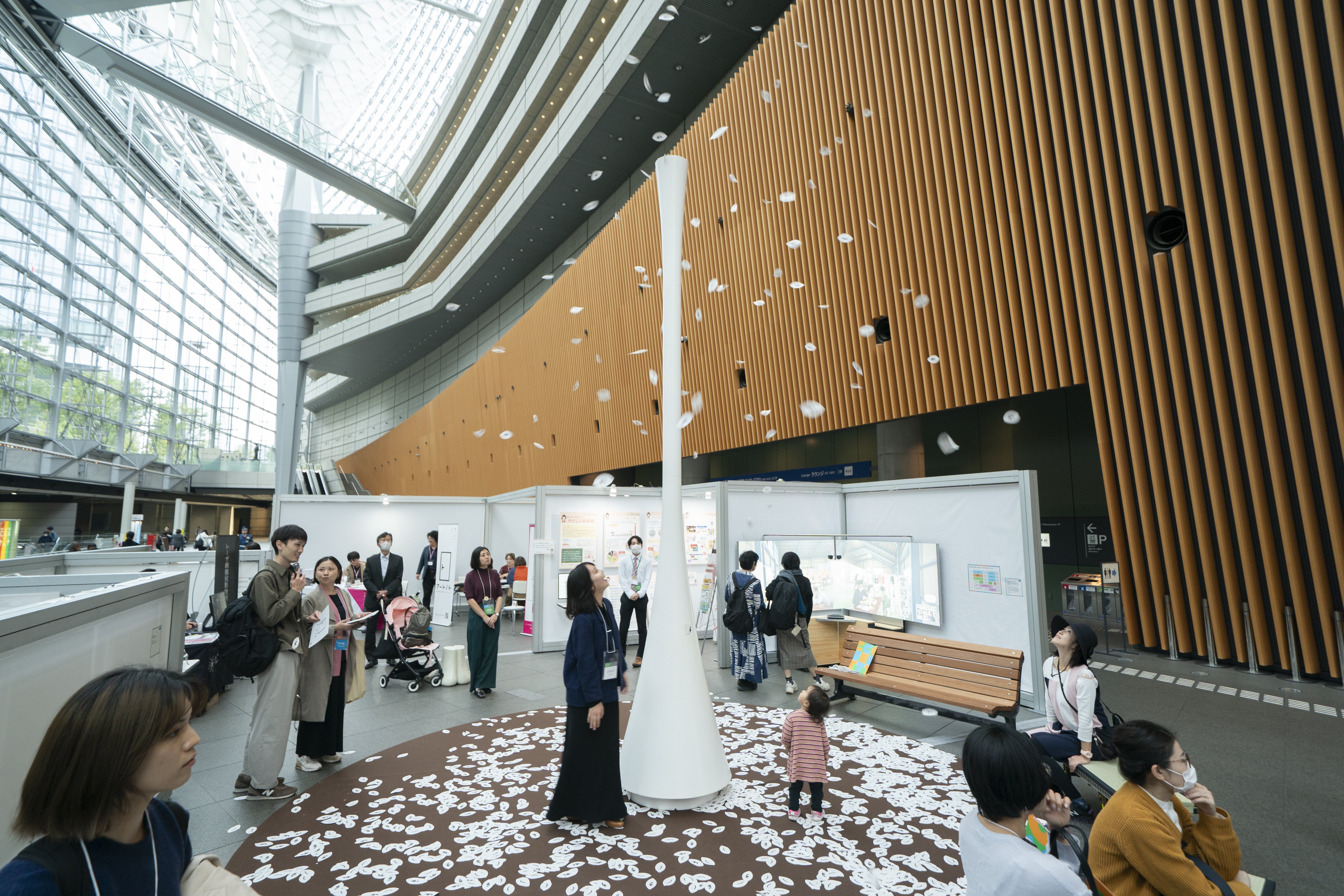 天井が高く、大規模な展示会場の風景。右側にはオレンジ色の湾曲した木製パネルの壁がある。中央には白く細長い円錐状のオブジェクトがあり、オブジェの頂点から白い葉っぱのようなものがたくさん吹き出されていて、床にはそれらが敷き詰められている。その様子を、周囲にいる多くの参加者が見つめている。