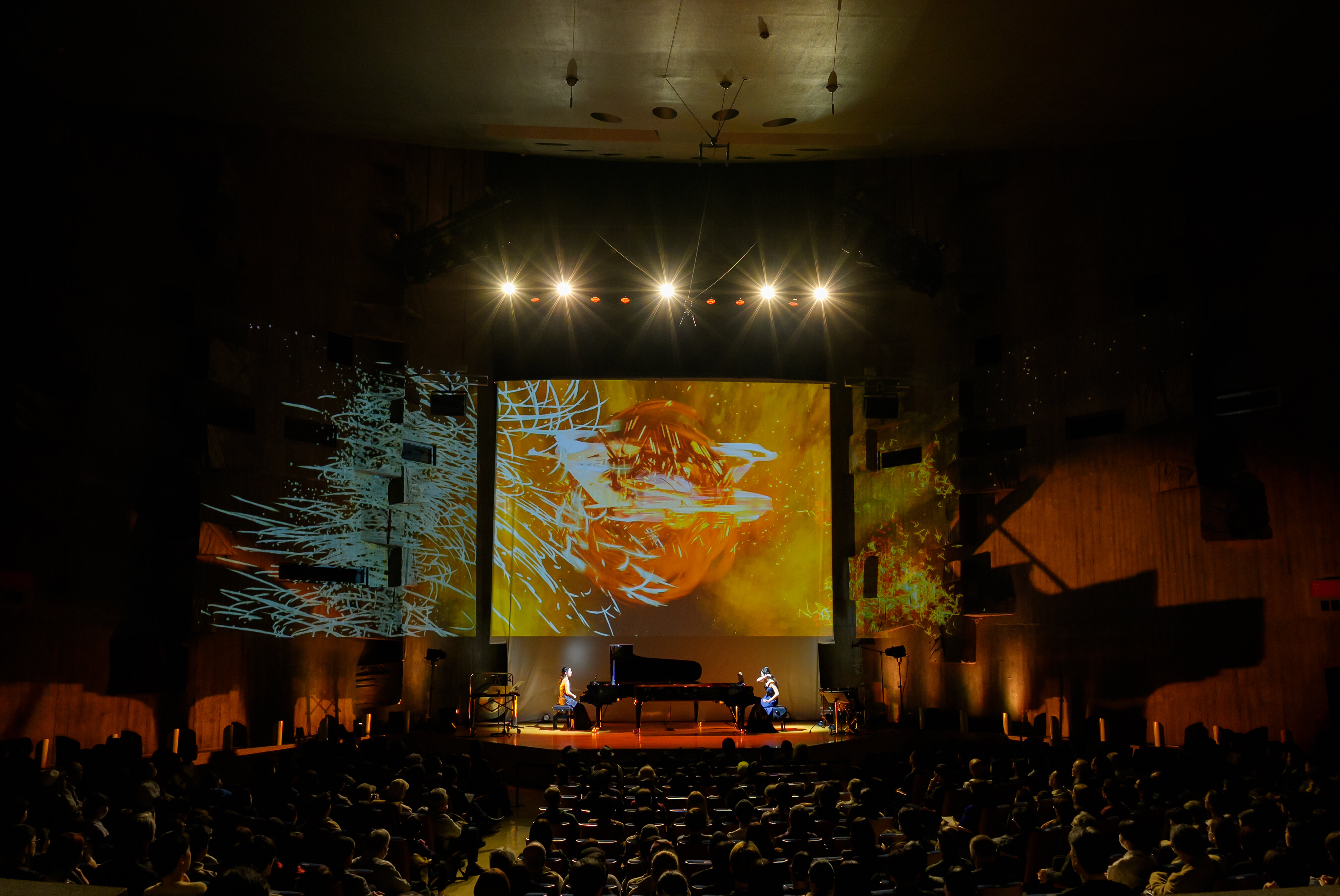大型モニターに火花が散っているようなダイナミックな映像ともに、舞台上で2人の女性が向かい合ってピアノデュオで演奏している様子