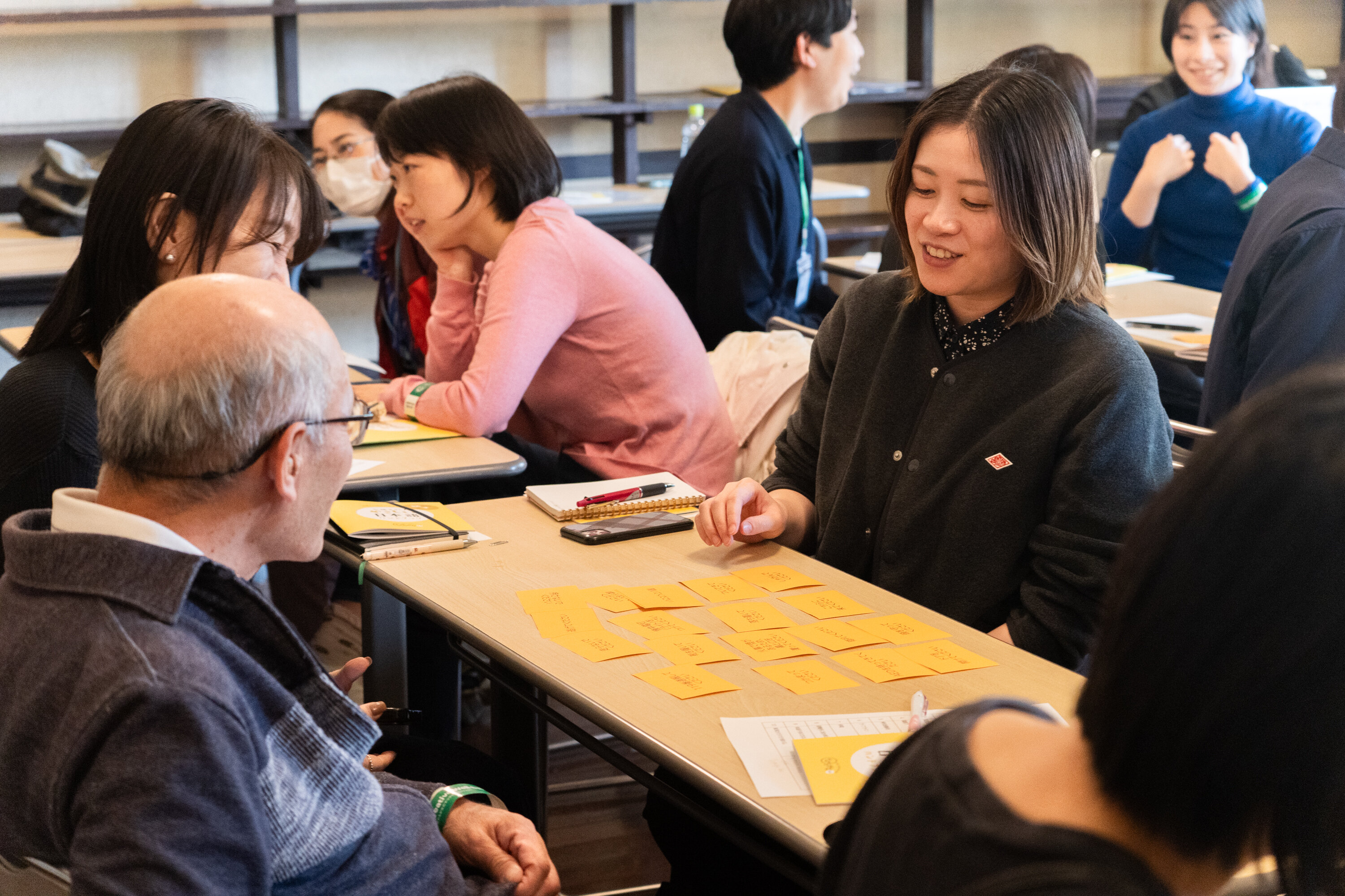 長机を囲むように８名の参加者が座っている。机の上には付箋が沢山貼られており、中央に座っている一人の参加者が付箋を見て、少し微笑みながら周りの参加者に何かを話している。