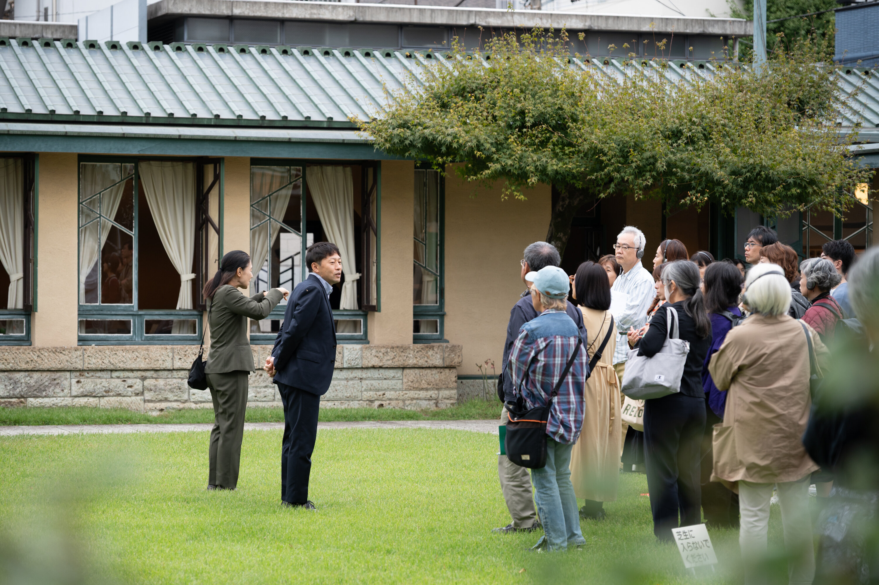 芝生の上で紺色のスーツを着た人が15名ほどの参加者に向かって説明している様子。その人の隣にはグレーのスーツを着た人が手話で通訳をしている。背後に、自由学園明日館の建物の一部が映っている。