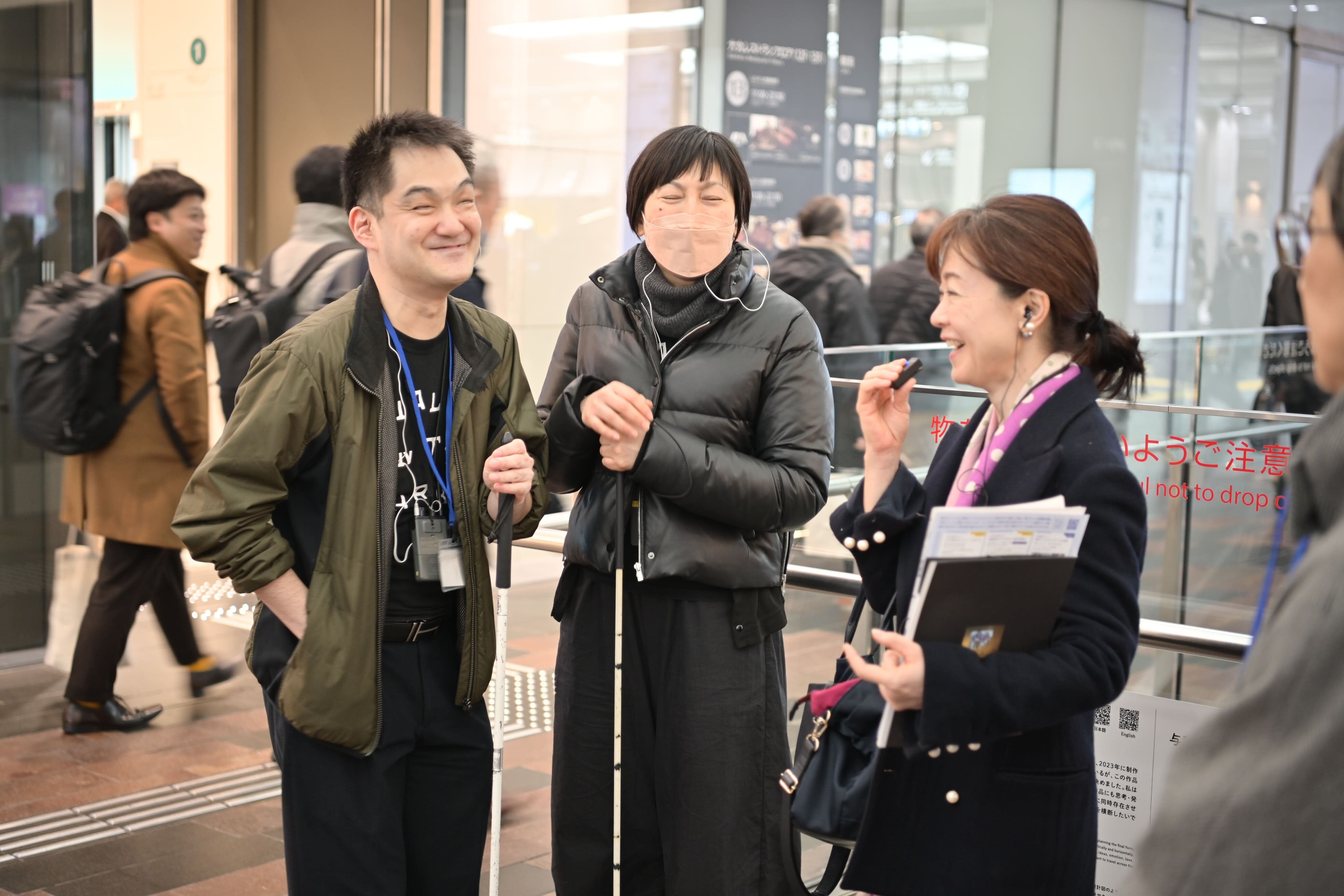 白杖を持っている2人の男女とそのとなりに小さなマイクのようなものを持って何か話している女性の写真