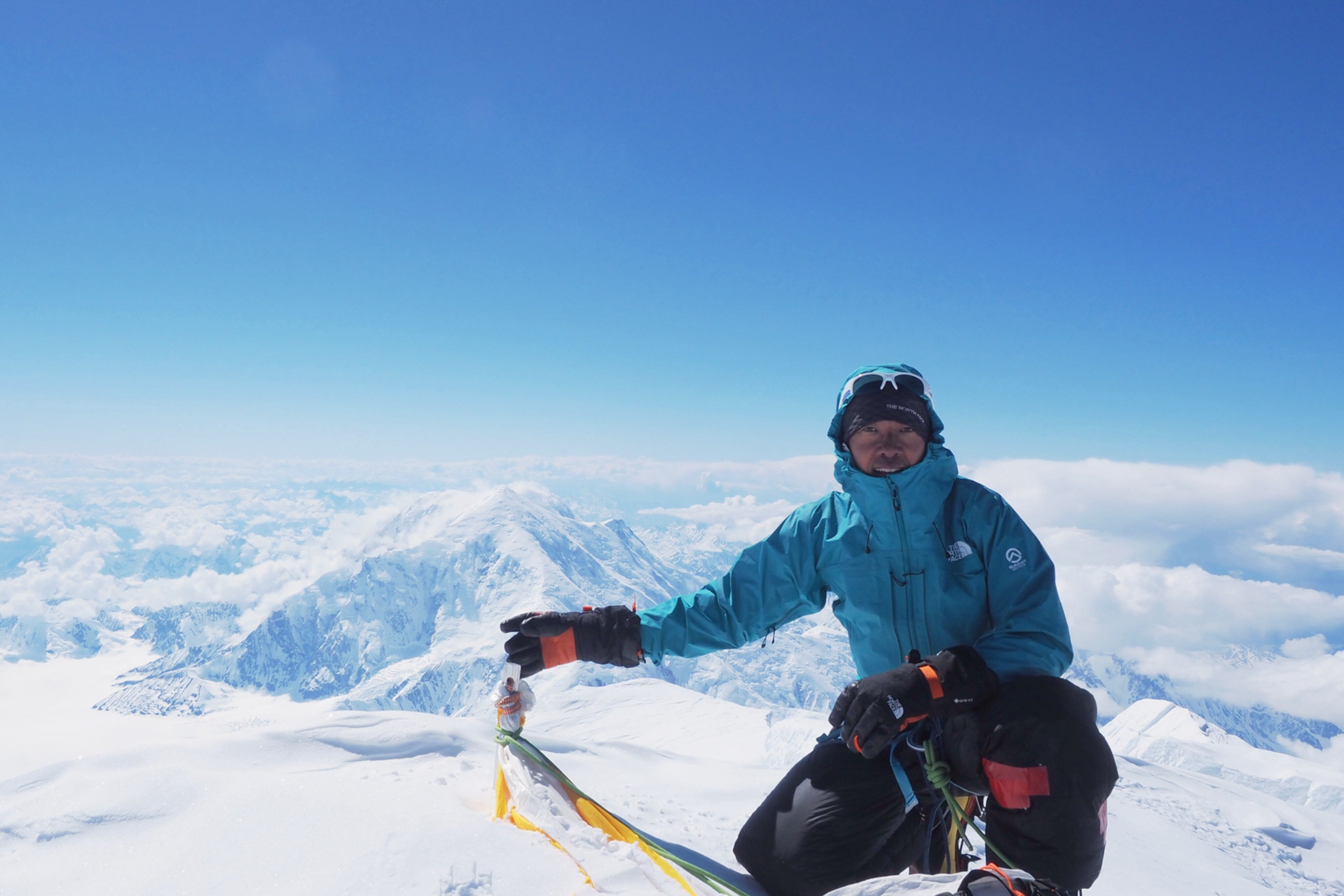 雪と氷に覆われた高山の頂上付近で、青空を背景に登山者の男性が片膝をついてカメラに向かってポーズをとっている。ブルーのジャケット、サングラスをヘッドバンドの上に装着し、黒い手袋とハーネス、ロープを身に着けている。背後には雪を被った山々の稜線と雲海が広がり、足元には目印の旗やロープが見える。