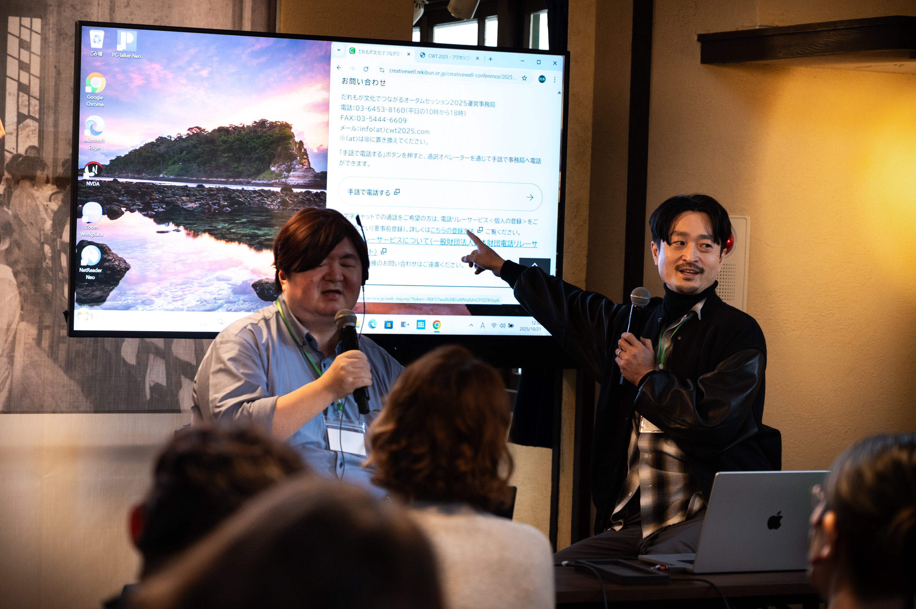 ２人の登壇者が手前にいる参加者に向かって話している。右側にいる登壇者がマイクを持ちながら背後のモニターのスライドを指差している。左側にいる登壇者は右側にいる人の説明を聞きながら何かを話している。