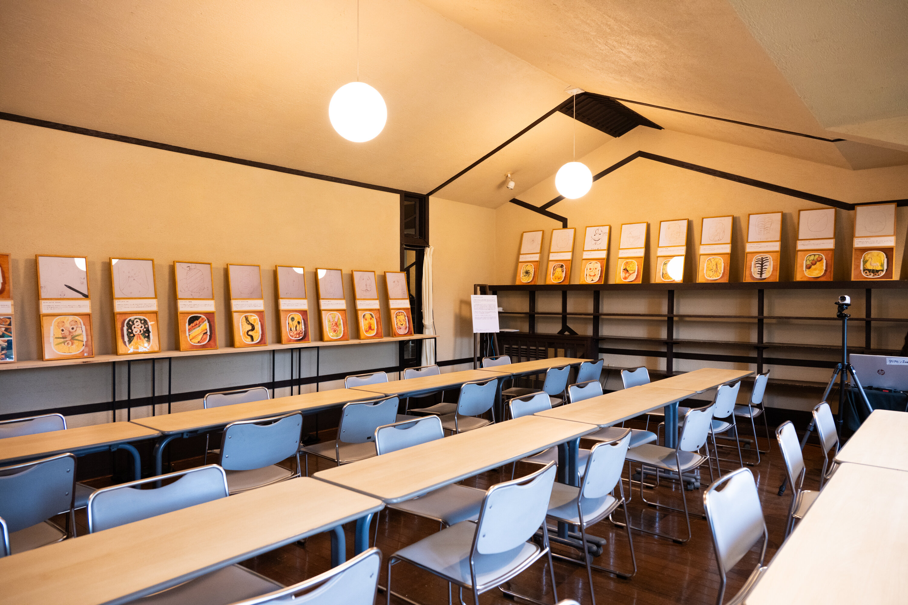 ２つの丸い照明が照らしている部屋の中に長机とイスが３列に並べられている。壁には、縦長の写真パネルが１５枚ほどズラリと飾られている。