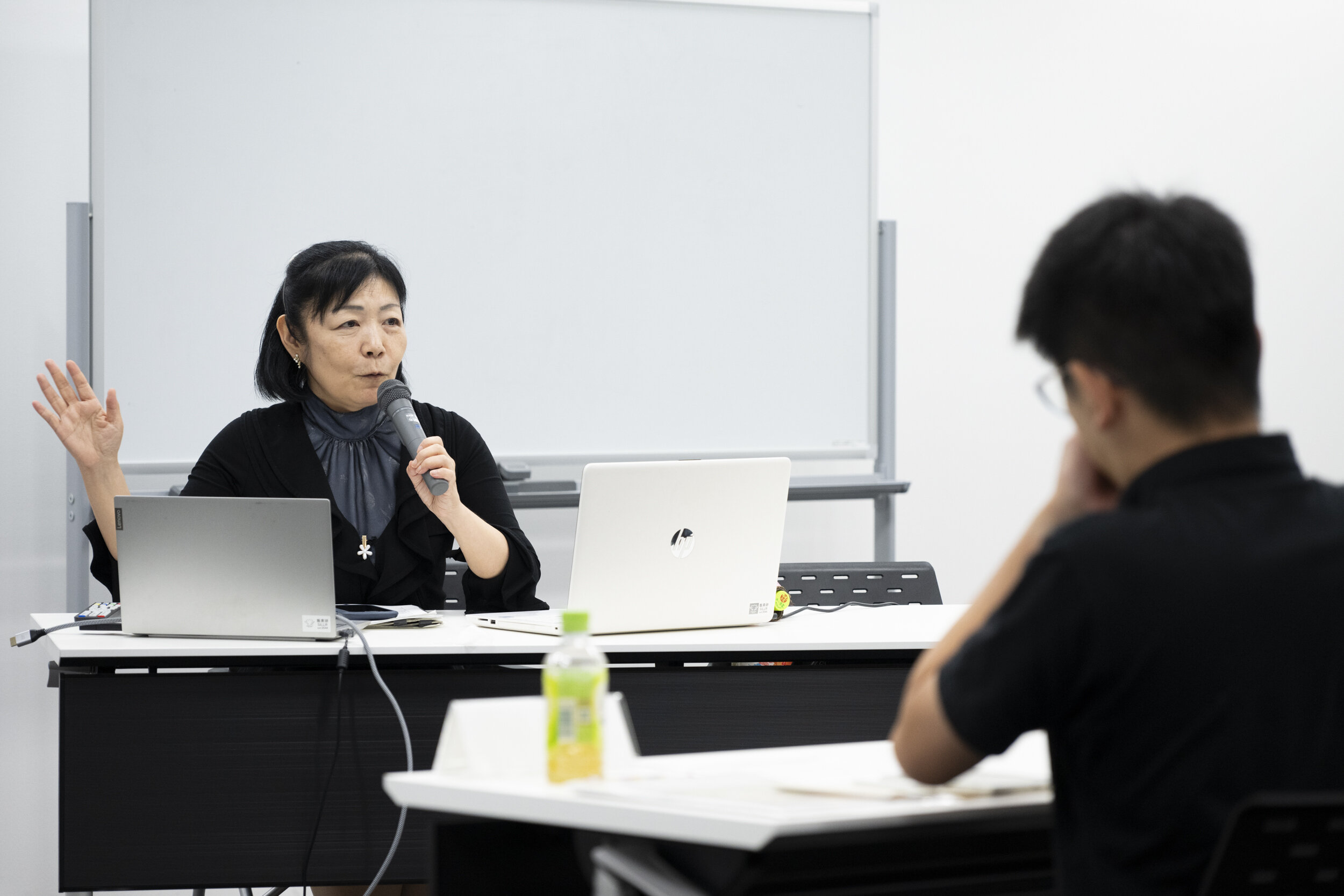 講師が椅子に座っており、机には2台のパソコンがある。講師がマイクを持って、目の前に座っている受講生に向かって話している様子の写真。