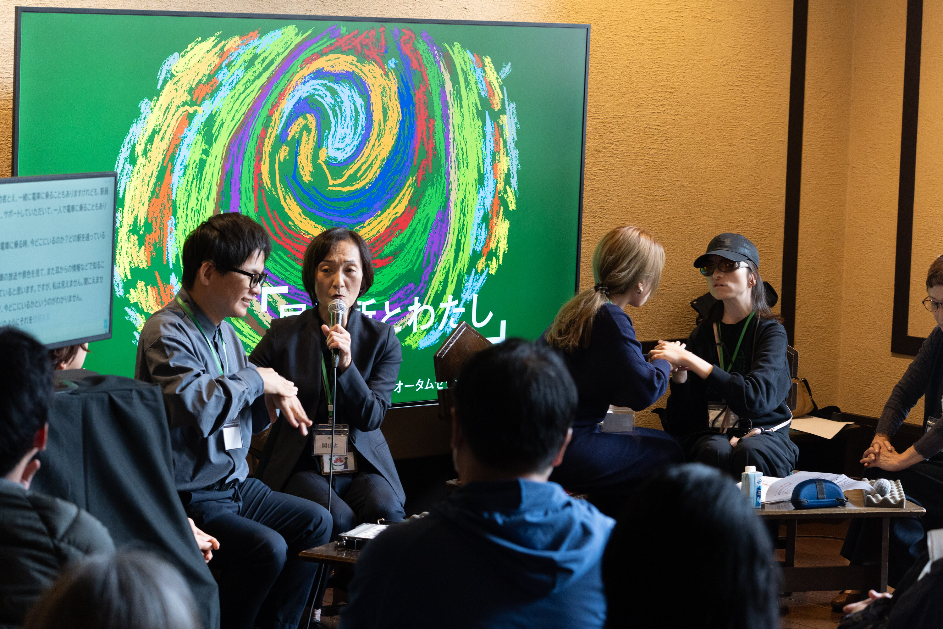 大きなスクリーンを背に、４人が座っている。一番左側の人はメガネをかけており手話で何かを話している。右隣の人が、その手話を見ながらマイクを持って説明している。一番右側の人はサングラスに帽子をかぶっており、その左隣にいる金髪の人の手話を手で覆うような形で触れている。
