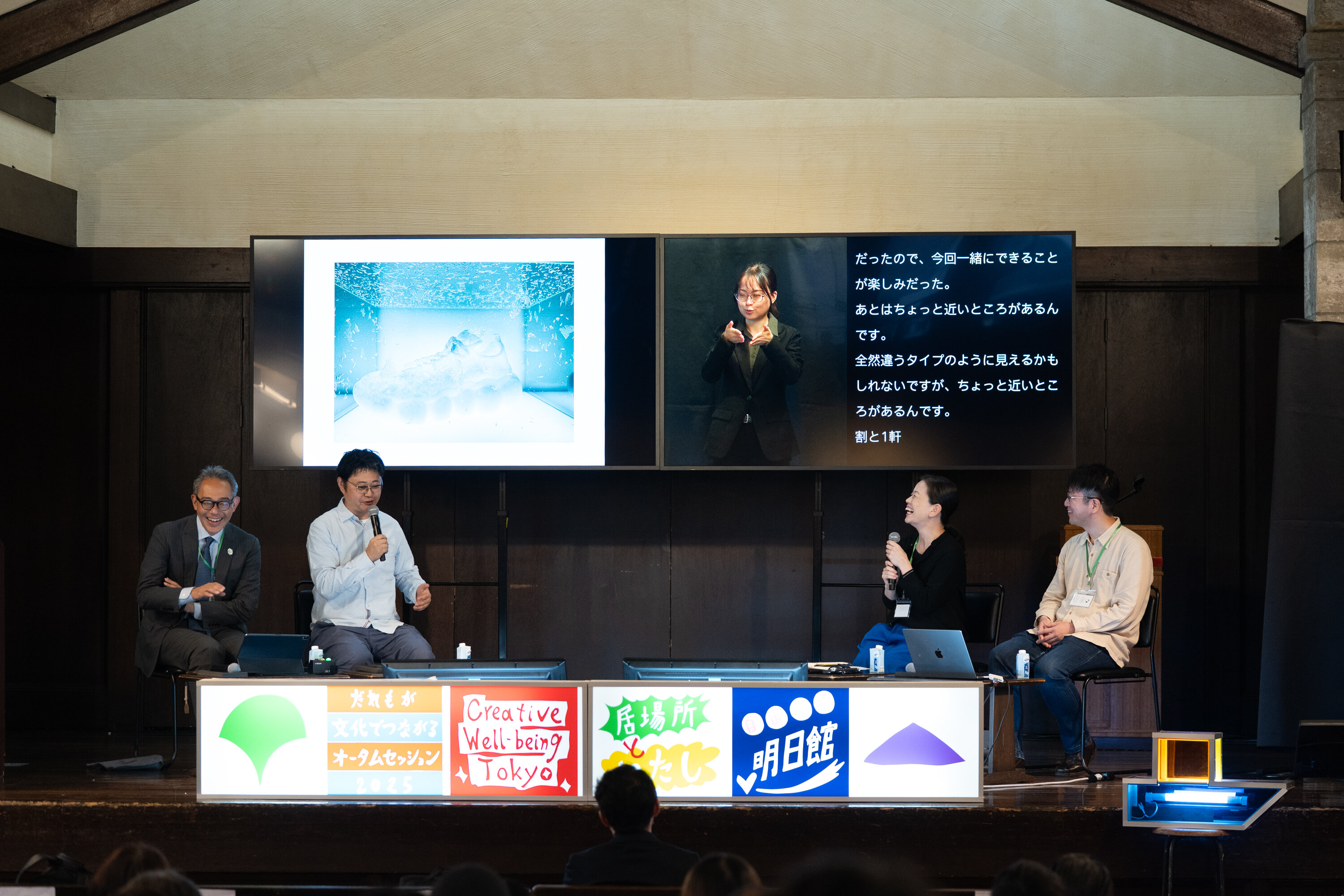 講堂内の舞台上の様子を中央から撮った写真。舞台中央に２つの大きなモニターが横に並べられている。右側のモニターには手話通訳と字幕が映し出されている。モニターの下に、４名の登壇者が座っている。登壇者の手前には横長の看板作品が置かれている。