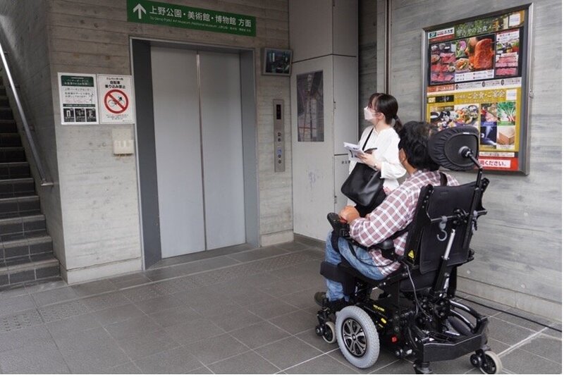 2名の調査スタッフが、エレベーターを待っている写真。一人は電動車椅子を使っている。エレベーターの脇の案内には、「上野公園・美術館・博物館方面」と書かれている。