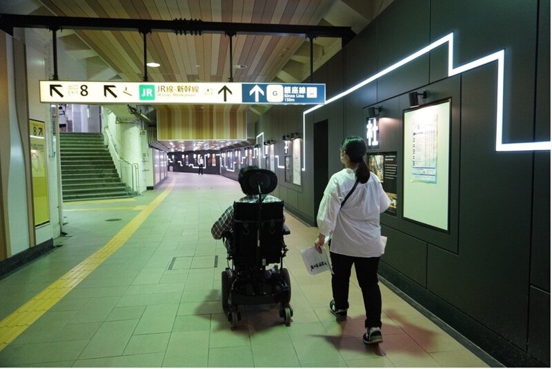 地下鉄の地下道を、2名の調査スタッフが歩いている写真。一人は電動車椅子を使っている。進行方向に、地下道の案内板や階段がある