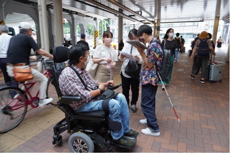 上野駅前で、白杖を使う人と電動車椅子を使う人を含んだ4名の調査スタッフが資料を見ながら話し合っている