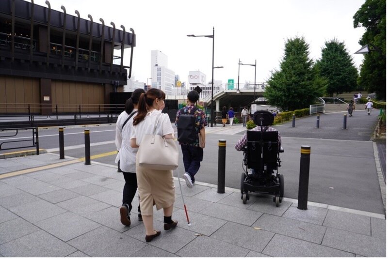 駅から文化施設への道のりの調査のために、電動車椅子を使う人と白杖を使う人を含んだ4名が屋外を歩いている写真