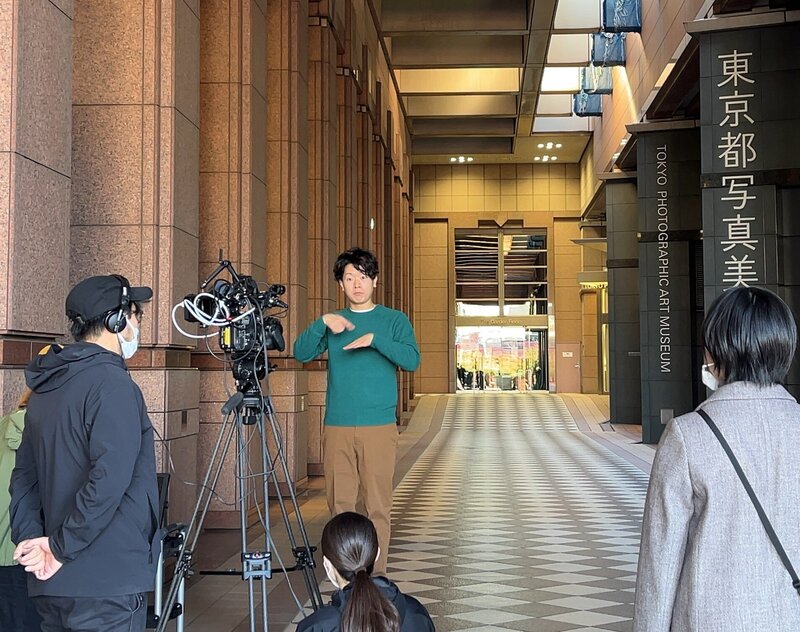 写真美術館で案内動画の撮影をしている写真映像のナビゲーターが写真美術館の入り口前に立って、カメラに向かって手話で説明をしている。