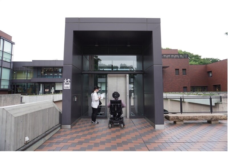 東京都美術館のエントランス階に降りるエレベーターを二人の調がスタッフのが待っている様子。一人は電動車椅子を使っていて、もう一人は紙とペンを持って何かをメモしている。