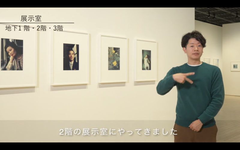 映像のスクリーンショット。展示室にいるナビゲーターが手話で展示室の説明をしている。画面の下部に日本語字幕で「2階の展示室にやってきました」と書かれている。
