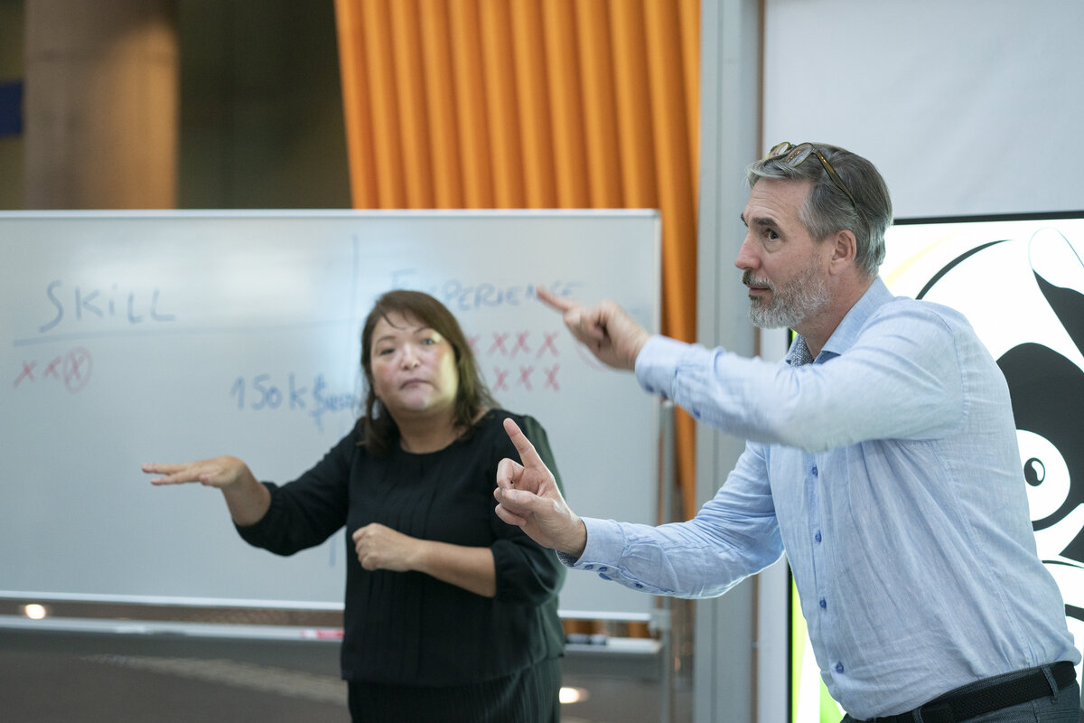 ホワイトボードを背に、デイビッド・デ・キーザーが手話で説明している様子。デイビッドの隣に手話通訳が通訳している。