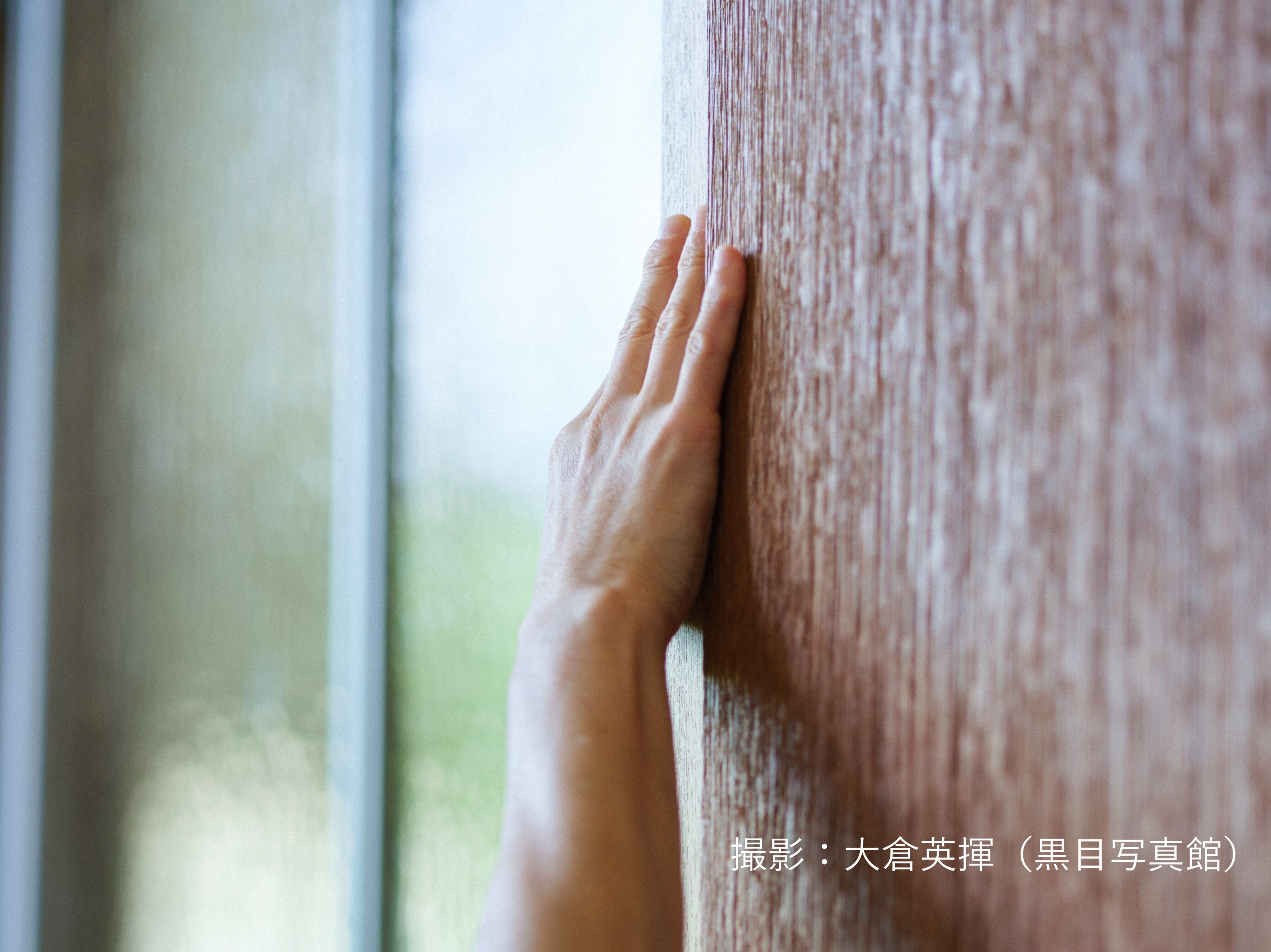 壁に右手を当てている写真。右下に撮影、大倉英揮（黒目写真館）と書いてある。