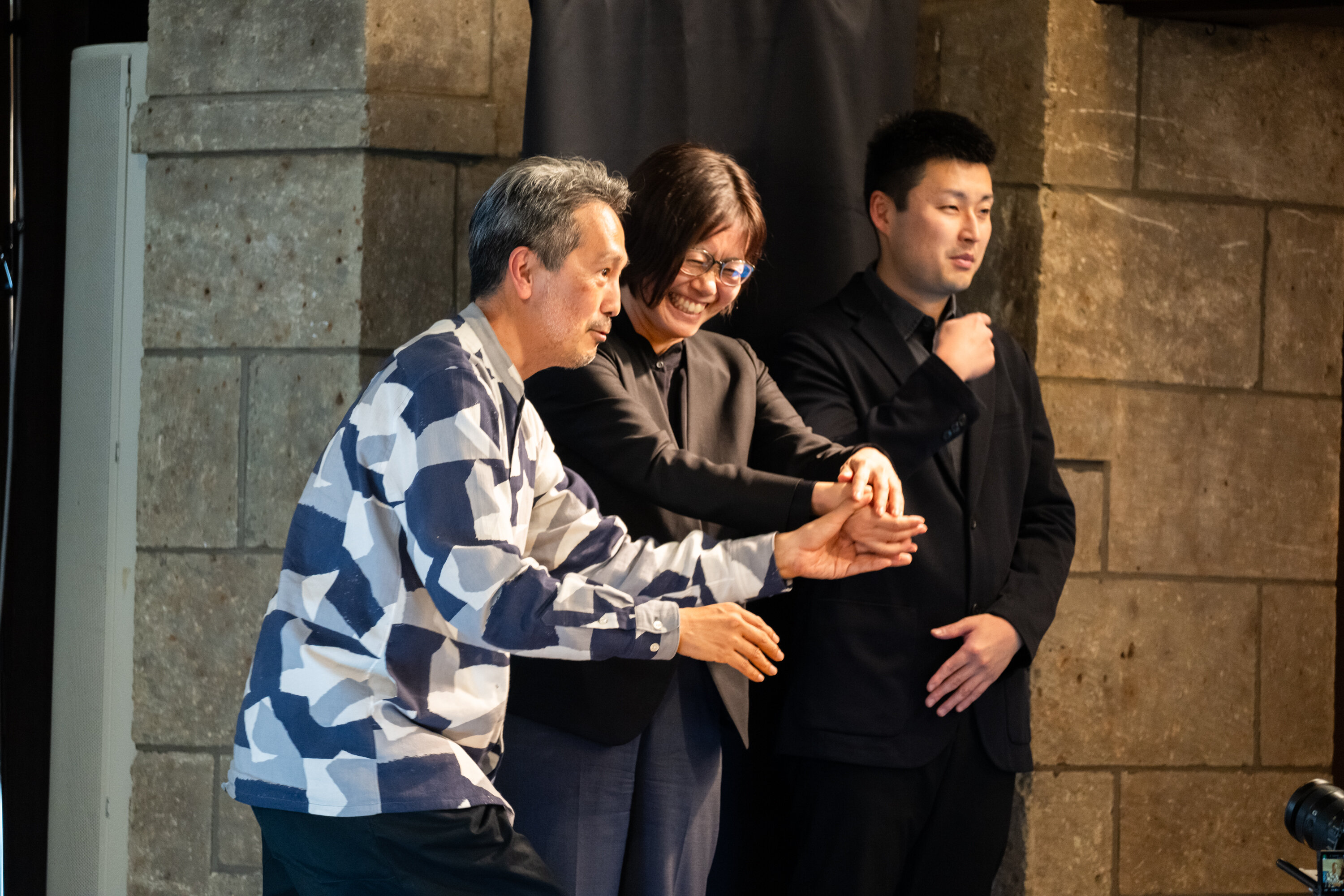 青とグレーのまだら模様の服を着た登壇者が眼鏡をかけた人の手を取り、ダンスのような動きをしている。眼鏡をかけた人は笑顔になっている。写真右側には手話通訳者が通訳をしている。