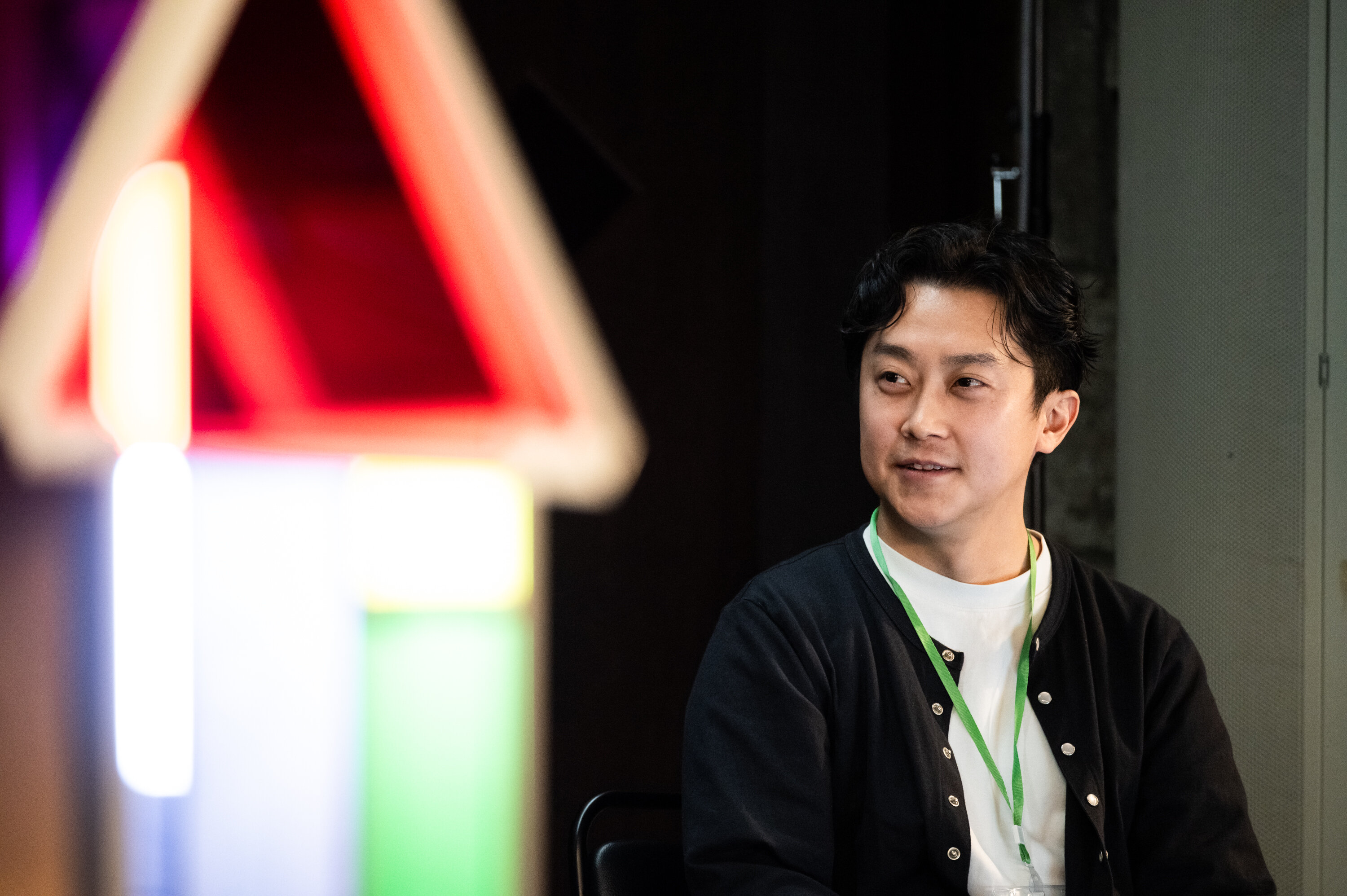 黒の上着に白のシャツを着た登壇者が左を見て微笑んでいる。写真の手前には家の形をしたカラフルなオブジェがぼんやりと映っている。