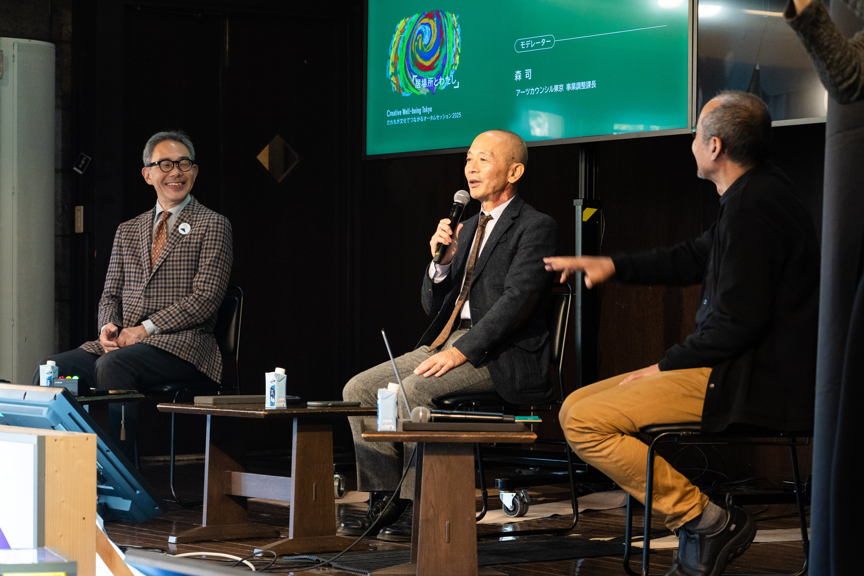舞台上に３名の登壇者が座っている。写真左側の眼鏡をかけた人は笑顔を見せ、中央の人はマイクを持って説明している。右側の人は手を前に出し、話に反応している。