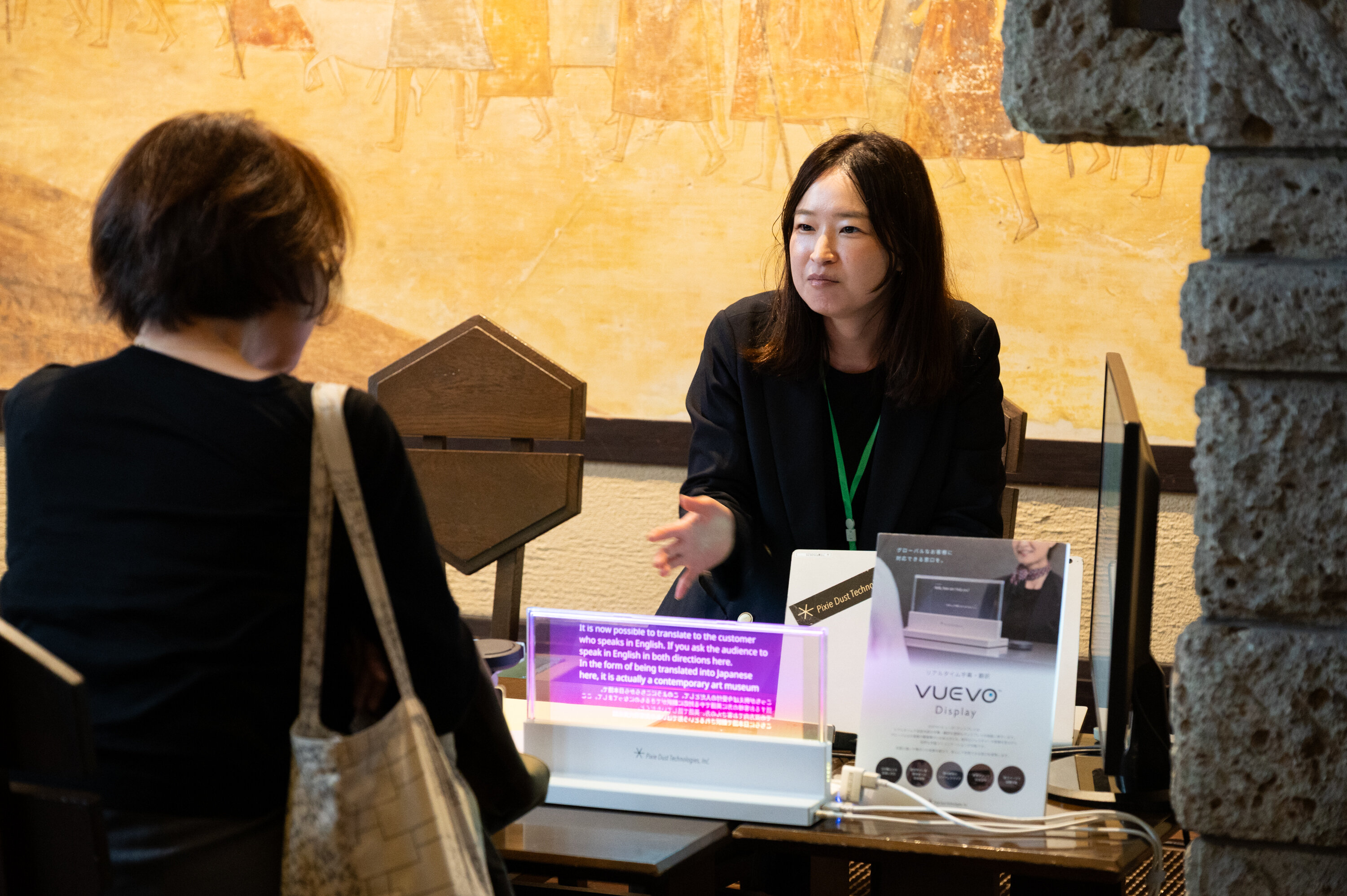 机をはさみ、2人が向かい合うような形で座っている。机の上には透明なディスプレイが置かれており、英語が表示されている。ディスプレイの隣には「VUEVO　ビューボ」と書かれているパネルが置かれている。奥に座っている人が手前に座っている人に、ディスプレイを手で示しながら説明している。