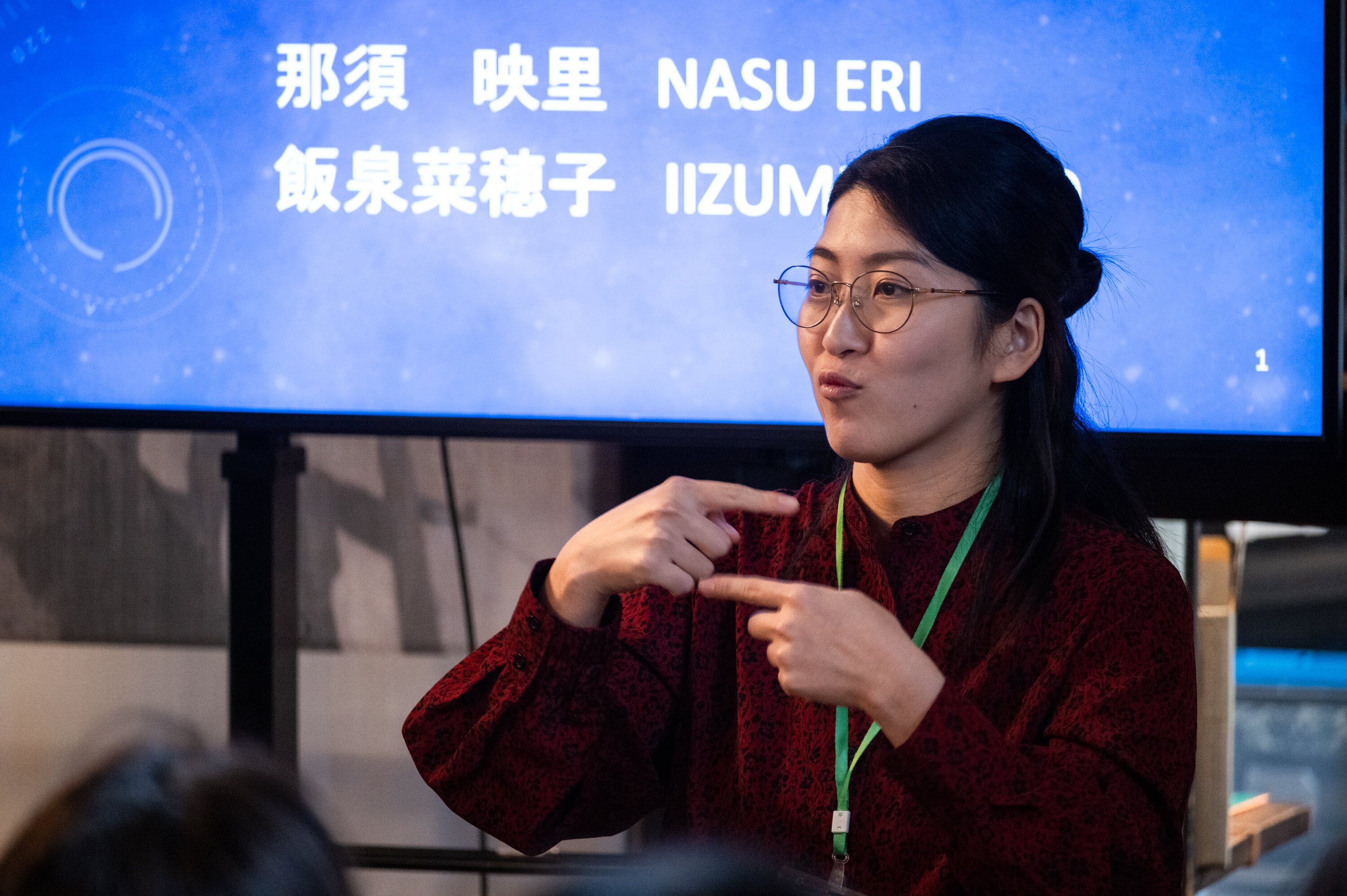 眼鏡をかけてえんじ色の服を着た登壇者が手話で何かを伝えている。背後のモニターにはスライドが投影されており、「那須映里」と「飯泉菜穂子」と書かれている。