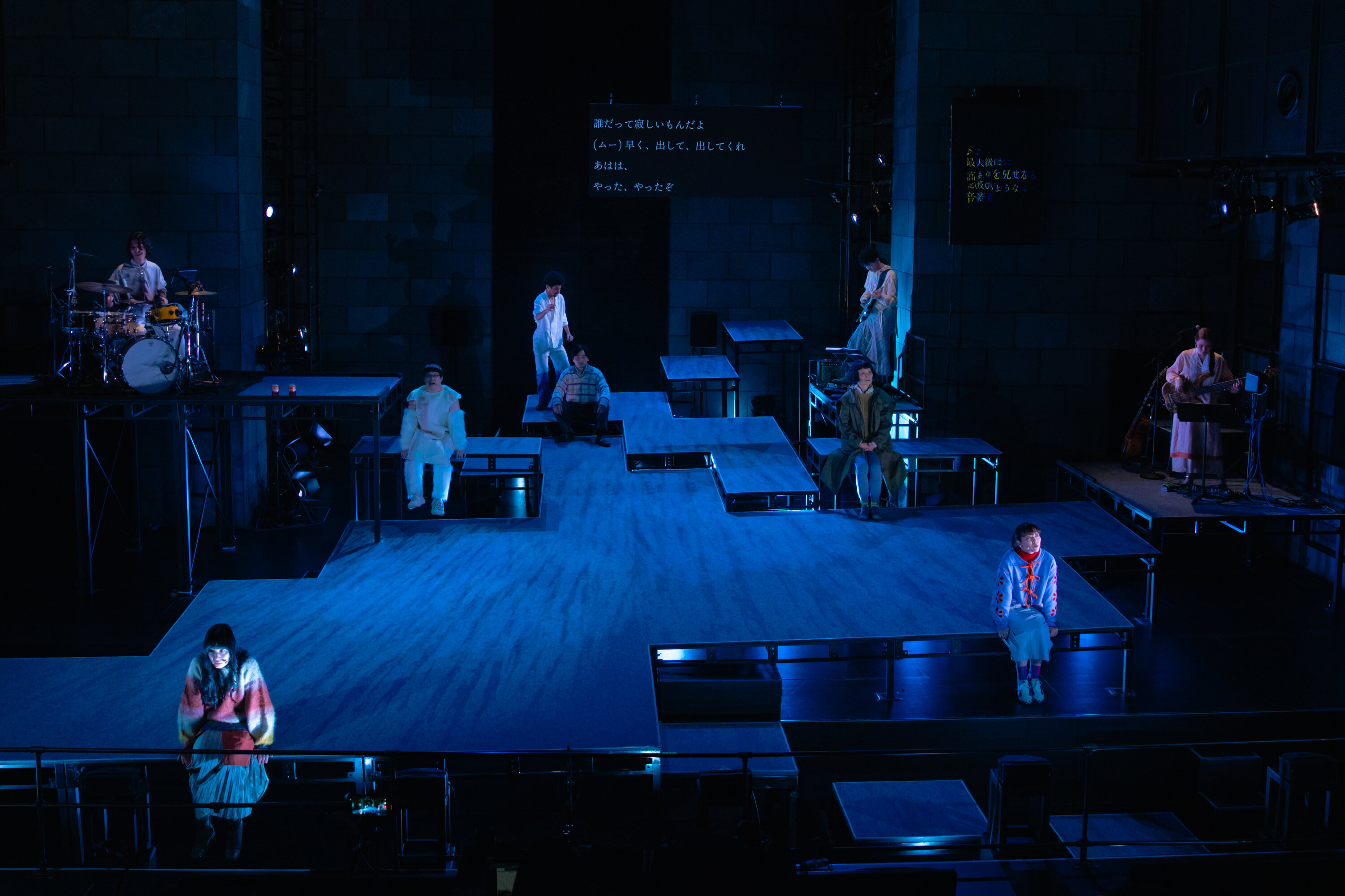 青暗い舞台の上で複数人が座ったり、演奏したりする様子の写真 