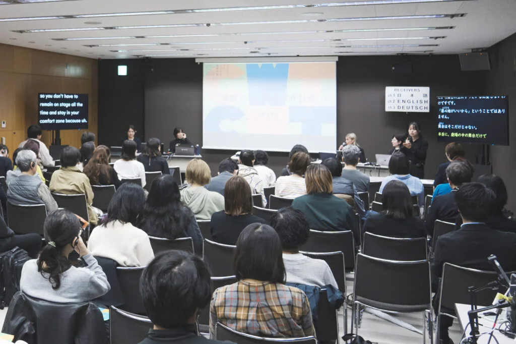 セミナーの写真。会議室のような部屋で手前には30名ほどの参加者が座っている。正面奥中央のスクリーンの前に4名の登壇者がこちらを向いて座っている。登壇者の左側 には英語の字幕モニター、右側には日本語の字幕モニターが設置されている。日本語の字幕モニターの隣に 手話通訳者が通訳している。