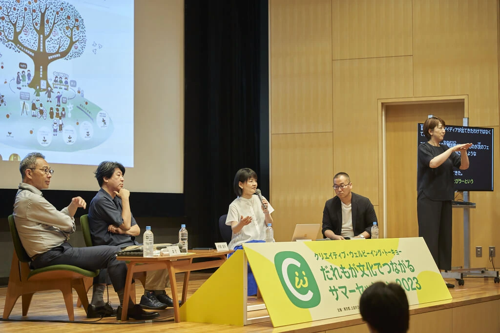 セッションの写真。ホールの壇上に4名の登壇者が観客に向かって座っている。そのうちの1名はマイクをもって何かを話している。壇上の右側には手話通訳が通訳しており、通訳の後ろには字幕モニターが置かれている。